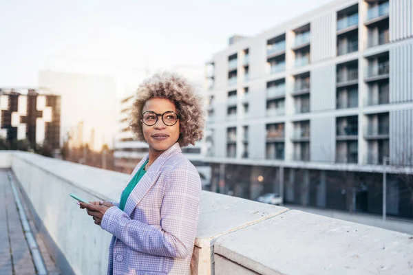 Professional black business woman with blond hair and trendy eyeglasses standing in the city looking away at sunset