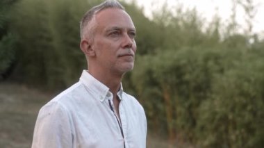 Olgun bir adamın portresi doğa ormanında duruyor, temiz hava soluyor, alacakaranlıkta doğayı düşünüyor. Orta yaşlı erkek, parkta sakin ve huzurlu bir hayatın tadını çıkarıyor. Mutluluk ve Memnuniyet 