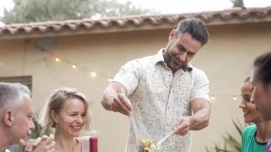 Mutlu İspanyol adam masaya sağlıklı makarna salatası servis ediyor. Arkadaşları akşam yemeği partisinde harika vakit geçiriyor.