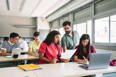 Dost canlısı profesör, lise öğrencilerine egzersizleri anlatıyor. Sakallı genç öğretmen genç kıza yardım ediyor.