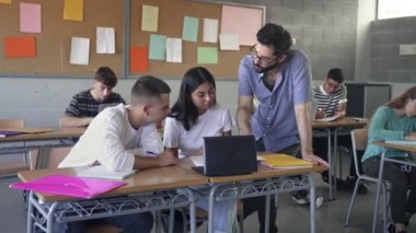 Young Professor assisting students in modern Secondary School classroom. Friendly Teacher with beard explaining assignment. Computer laptops and new technology in Education