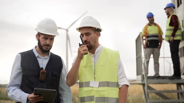 風力タービン発電所で働く電気技術者と技術者のチームは タブレットとウォーリートーキーを使用して技術検査を行う — ストック動画