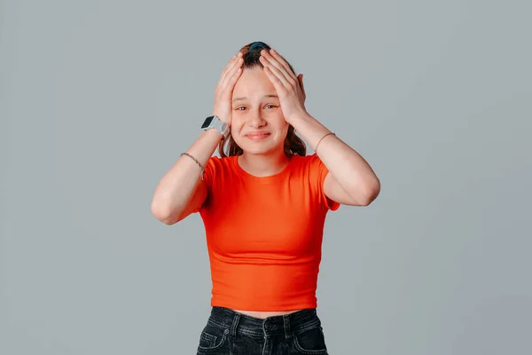 頭の上に手を握っているブルネットの十代の少女の肖像画は 良いニュースを聞いて 灰色の背景にオレンジ色のTシャツに立って 驚きと驚きを見ています 若い感情的な女性 — ストック写真