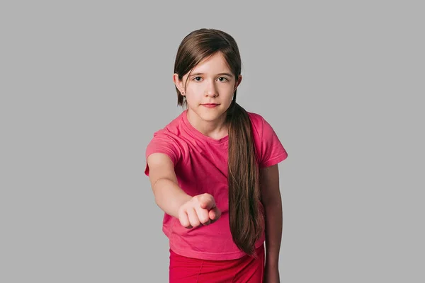 若い深刻な長い栗の厚さの髪を持つ女の子 カジュアルなピンクのTシャツを着て カメラに向かって指を指して 灰色の背景の上に立って — ストック写真
