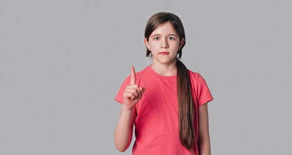 年轻而严肃的黑发女孩摇动着手指 指尖向上 看上去很自信 告诉人们注意 禁止涂抹 发出警告 穿着粉色T恤 站在灰色的背景上 复制空间 — 图库照片