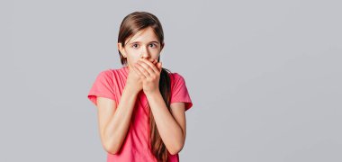 Uzun kestane rengi gür saçlı, korkmuş genç kız ellerini kameraya uzatarak ağzını kapadı. Gri arka planda duran günlük pembe tişörtünü giydi. Genç duygusal kadın.