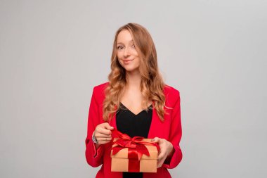 Kırmızı kurdeleli, hediye kutusu olan çekici bir genç kadın. Sarı kıvırcık saçlı, şık kırmızı ceketli, gri arka planda duran mutlu kadın. Yeni yıl kadınlarının doğum günü konsepti