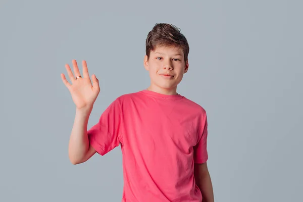 Dost canlısı genç çocuk el sallıyor ve merhaba diyor selam veriyor, seni gri arka planda pembe tişörtle görmek son derece mutlu ediyor.