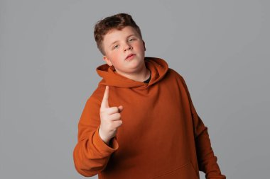 Ciddi kararlı genç çocuk reddedilmek için parmağını sallıyor, yasaklıyor ya da yasaklıyor, kötü davranışlar için azarlıyor, suratını asıyor ve hayal kırıklığına uğruyor, gri arka plan üzerinde sıradan giysiler içinde duruyor.