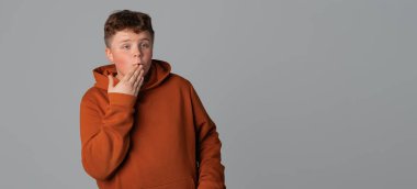 Tüh. Heyecanlı ve şaşırmış görünen genç bir çocuk görüntüsü, ağzını kapatan, ilginç söylentiler duyan, gri arka planda duran, metin için boş alan olan günlük giysiler.