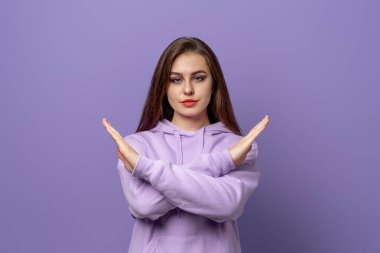 Uzun saçlı, ciddi genç bir kadın durgun bir jest yapıyor, eğlenmemiş görünüyor, hayır diyor, bir şey yasaklıyor, mor kapüşonlu giyiyor, mor arka planda duruyor.