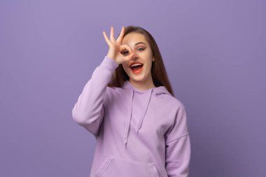 Gülümseyen esmer bir kadının mor arka planda mutlu bir yüz ifadesiyle iyi bir jestte bulunmasına sevindim. Kameraya bakıyorum. Pozitif duygu kavramı