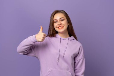 Mutlu gülümseyen, güzel, esmer bir kadın baş parmağını kaldırıyor, mor arka planda duruyor. Pozitif duygu kavramı