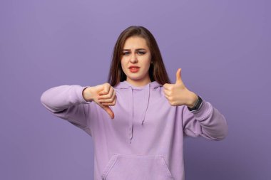 Güzel, genç, esmer bir kadının baş parmağını kaldırması ve baş parmağını kaldırması zor bir seçim, fikir ayrılığı ve anlaşma ifadesi, mor arka plana karşı durması. Çılgın çatışma