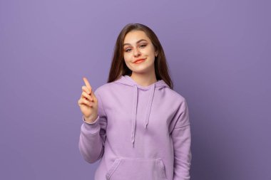 Kafası karışmış genç bir kadın işaret parmağını sallıyor ve suratını ekşitiyor, önerileri ya da endişeleri var, görüşlerini söylüyor, garip durumları yargılıyor, mor arka planda modaya uygun bir kapüşonluyla duruyor.