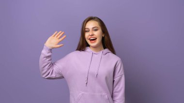 Dost canlısı, neşeli, genç esmer kadın gülümseyen, selam veren ve el sallayan kişi, merhaba hareketi yapan, hoş geldin diyen, mor arka planda modaya uygun kapüşonlu biri.