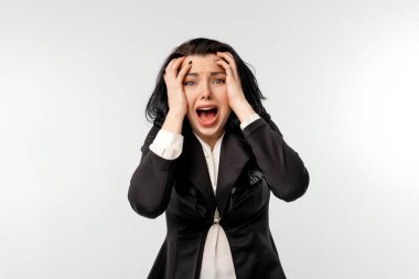 Paniklemiş iş kadını, siyah ceketli, beyaz gömlekli, kafasına yapışan. Şaşırmış genç iş kadınının portresi kameraya bakıyor ve beyaz odada tek başına ağzını açık tutuyor.