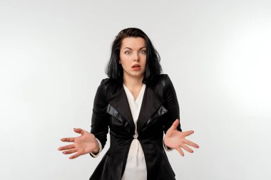 Beyaz arka planda siyah ceketli, beyaz gömlekli, sürpriz bir iş kadını. Ağız açık, ellerini yana açıyor, şok olmuş bir suratla kameraya bakıyor. İş konsepti