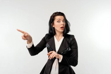 Şok olmuş iş kadını, duyuru yapıyor, solu işaret ediyor, siyah resmi bir ceket giyiyor, beyaz arka planda dikiliyor.