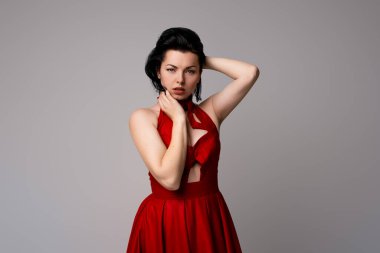 Gri arka planda duran, kırmızı elbiseli, zarif, koyu renk saçlı genç bir kadın.