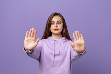 Ciddi genç esmer kadın dur işareti yapıyor, hayır diyor, bir şeyi yasaklıyor, mor kapüşonlu giyiyor, mor arka planda duruyor. Evde kal. Sosyal mesafeler. Yeni normal davranışlar.