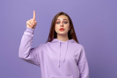 Uzun saçlı güzel bir kadın, parmağını kaldırıyor, kaşlarını kaldırıyor, şaşırıyor, reklamını yapıyor, mor arka planda duruyor.