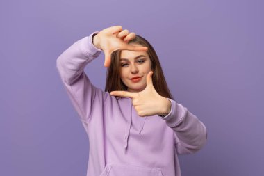 Mutlu öğrenci kız mor arka planda elleri ve parmakları ile çerçeve yapıyor. Kameraya bakıyorum. Pozitif duygu kavramı