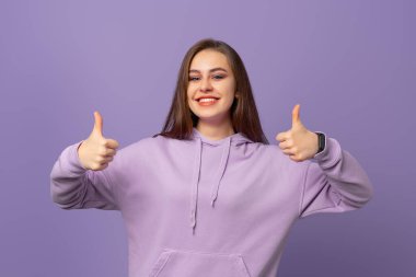 Gülümseyen güzel genç bir kadın onaylama jesti yapıyor ve kabul ediyor, mor arka planda duruyor. Pozitif duygu kavramı