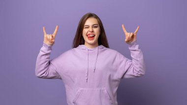 Sevinçli duygusal kız sevinçle bağırıyor, iki eliyle rock 'n roll hareketi yapıyor, en sevdiği yüksek sesli müzikten hoşlanıyor, mor arka planda duruyor. Bu partiyi salla.