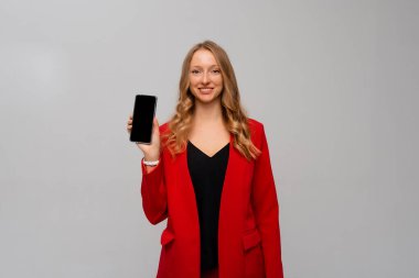 Güzel ve kendine güvenen iş kadını akıllı telefon ekranı gösteriyor, mobil uygulama gösteriyor, gri arka plana karşı kırmızı ceketle duruyor.