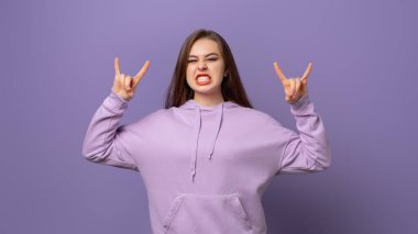 Duygusal esmer kız rock 'n roll hareketi yapıyor, bağırıyor, aşırı sevinç duyuyor, müzik temalı partiden hoşlanıyor, moda kapüşonlu giyiyor, mor arka planda izole edilmiş.