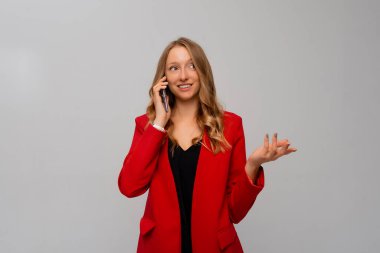 Güzel, gülümseyen bir iş kadını cep telefonuyla konuşuyor, bir telefona gülüyor, gri arka planda kırmızı ceketle dikiliyor. İş ve teknoloji