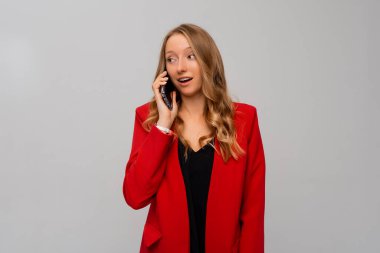 Güzel, şaşkın sarışın bir kadının cep telefonuyla konuşması, cep telefonuyla araması, gri arka planda kırmızı ceketle durması.
