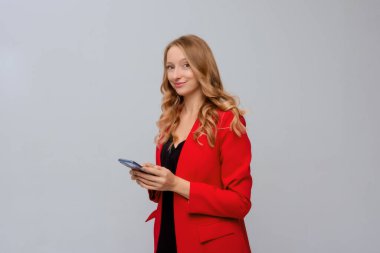 Başarılı genç girişimci kadın portresi, elini cep telefonuyla tutuyor, cep telefonunda gülümsüyor ve daktilo kullanıyor, mutlu bir yüz ifadesiyle kameraya bakıyor, gri arka planda duruyor.