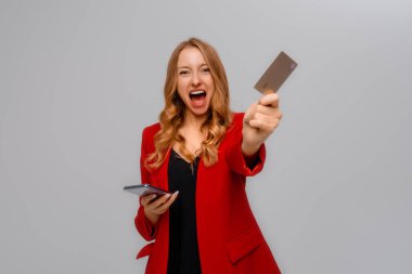 Mutlu bir şirket kadını, elinde akıllı telefon ve kredi kartı tutuyor, mağaza satışı için heyecanlı görünüyor, internetten ödeme yapıyor, cep telefonu siparişi veriyor, başarılı bir satın alma sayesinde mutlu bir şekilde bağırıyor, gri bir geçmişi var.