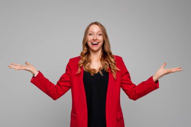 Ne olmuş, kimin umurunda. Mutlu gülümseyen genç sarışın kadın belirsizlik jestini sorgulamak için omuzlarını silkiyor ve avuç içlerini açıyor, gri arka planda kırmızı ceketle dikiliyor.