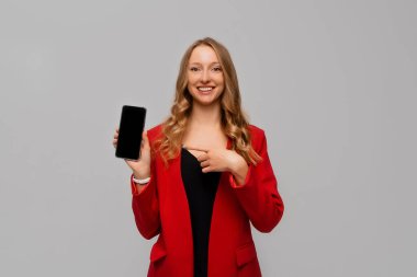 Business and technology. Smiling beautiful and confident business woman pointing finger at smartphone screen, showing app interface, standing in red blazer against gray background