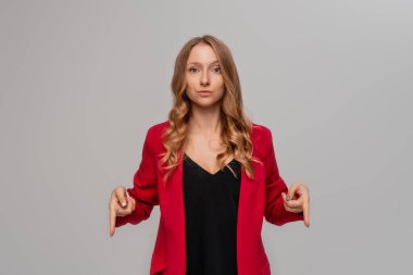 Skeptical blonde young woman pointing fingers to the down, looking seriously, showing something interesting, stands in red blazer against gray background