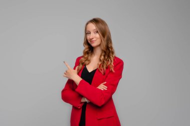 confident young woman points finger at upper left corner, showing to the advertisement, looking determined at camera, standing in red blazer against gray background