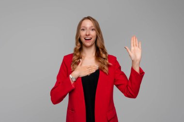 Smiling business woman introduce herself, raising hand and holding palm on heart, making promise, looking happy, standing in red blazer against gray background