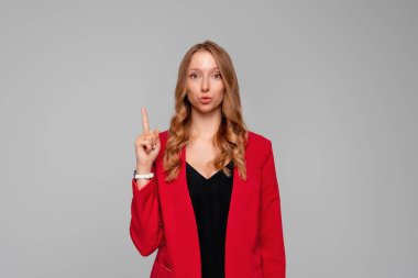 Serious female entrepreneur with fair hair in red and black clothes pointing finger up, scolding or telling off someone, says: Attention please, gray background