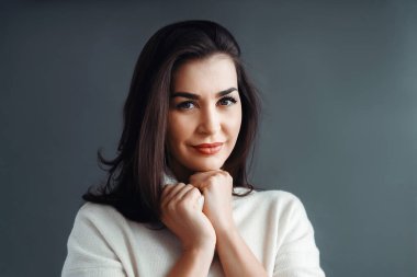 A beautiful brunette woman in a white sweater poses on a gray background in the studio. Beauty and elegance.