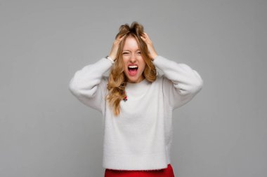 Stress. Woman stressed is going crazy pulling her hair in frustration and shouting from regret. Young emotional woman, wears white sweater, standing over gray background