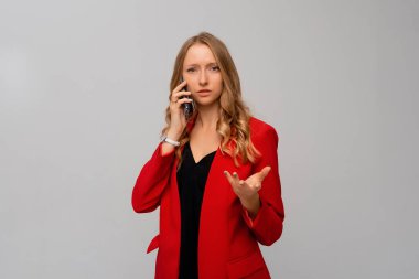 Confused young woman having a strange phone call, shrugging and looking puzzled, talking on mobile, standing in red blazer against gray background