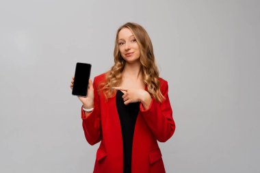 Business and technology. Beautiful and confident business woman pointing finger at smartphone screen, showing app interface, standing in red blazer against gray background