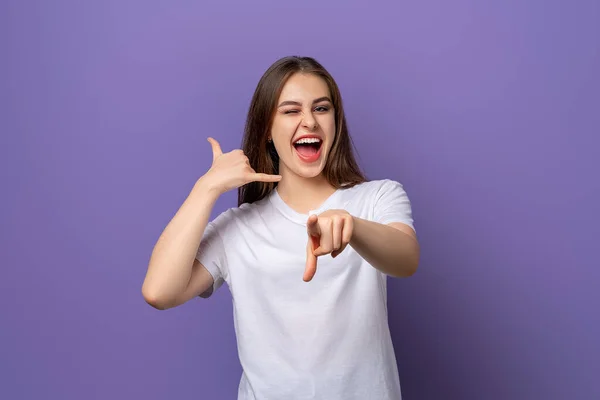 Portrait of smiling brunette girl pointing finger at camera, showing mobile phone gesture, call me, standing in blank white t shirt over purple background, standing over white background