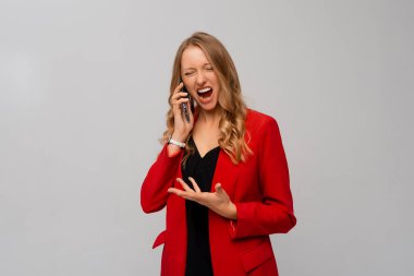 Kızgın genç iş kadını agresif bir şekilde bağırıyor, çok kızgın, hayal kırıklığına uğramış ya da sinirlenmiş görünüyor, hayır diye bağırıyor ve elinde bir cep telefonu, gri arka planda kırmızı ceketle duruyor.