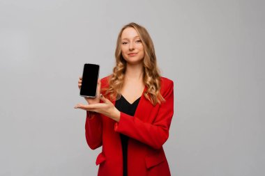 Güzel ve kendine güvenen iş kadını akıllı telefon ekranı gösteriyor, mobil uygulama gösteriyor, gri arka plana karşı kırmızı ceketle duruyor.