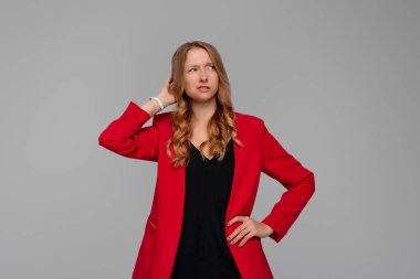 Tereddütlü, düşünceli, genç, sarışın bir kadın şaşkın görünüyor, bir şey hakkında düşünüyor, gri arka planda kırmızı ceketli duruyor. Kafası karışmış kadın