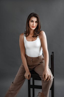 Tasasız genç bir kadın, gündelik kıyafetleriyle bir sandalyede oturuyor, stüdyoda gri arka planda poz veriyor. Rahat ve güzel bir genç kadın.